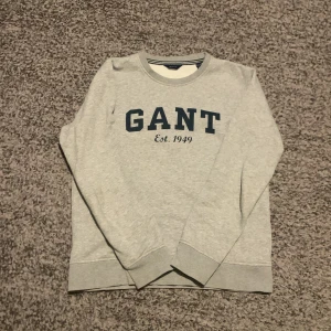 Grå sweatshirt från GANT - Säljer en stilren grå sweatshirt från GANT storlek 152 med mörkblå text 'GANT Est. 1949' på framsidan. Tröjan har långa ärmar och en rund halsringning med ribbade muddar vid ärmslut och nederkant. Perfekt för en avslappnad stil.S