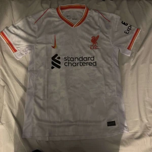 Liverpool 24/25 fotbollströja  - Säljer en vit fotbollströja från Nike med röda och gula detaljer. Tröjan har korta ärmar och klubbens logga på bröstet. Perfekt för fans som vill visa sitt stöd.
