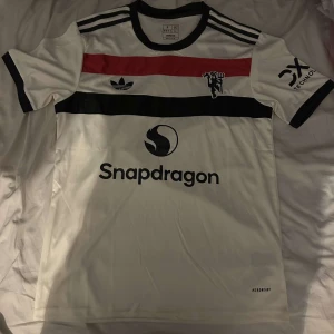 Manchester United 24/25 fotbollströja - Säljer en vit fotbollströja från Adidas med svarta och röda detaljer. Tröjan har korta ärmar och en svart krage. Den har logotyper och text på framsidan samt tre svarta ränder på axlarna. Perfekt för fotbollsfans! ⚽