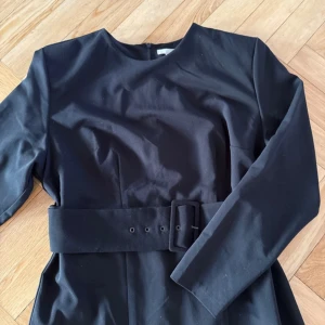 Svart klänning med bälte - Elegant svart klänning med långärmad design och ett matchande bälte i midjan. Perfekt för en stilren look. Små axelvaddar. Använd en gång. 