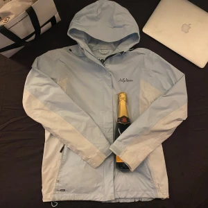 Ljusblå vindjacka från Helly Hansen - Säljer en ljusblå vindjacka från Helly Hansen med huva och dragkedja. Jackan har en stilren design med justerbara detaljer och är perfekt för blåsiga dagar. Passar bra för utomhusaktiviteter och har en praktisk ficka framtill.