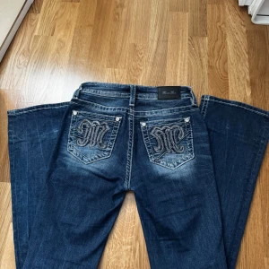 Mörkblå jeans från Miss Me - Snygga mörkblå jeans från Miss Me med dekorativa bakfickor och kontrastsömmar. Jeansen har en bootcut-stil och är perfekta för en avslappnad look. De har en knapp och dragkedja framtill. De har ett hål på ena benets baksida