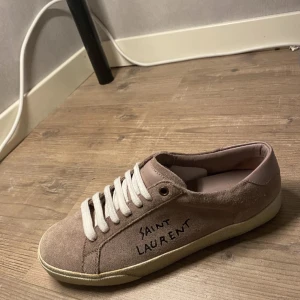 Beige mocka sneakers från Saint Laurent - Snygga beige sneakers i mocka från Saint Laurent med vit snörning och handskriven logga på sidan. Perfekta för en stilren look. Knappt änvända och i mycket bra skick storlek 40 men är stor i storlek så passar 41 också. Prisk kan diskuteras. Har även äktighetsbevis