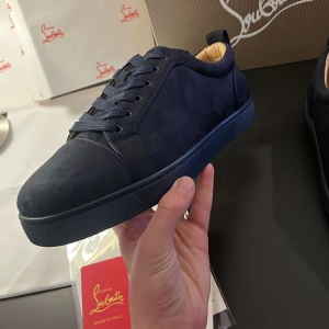 Mörkblå sneakers från Christian Louboutin - Snygga mörkblå sneakers från Christian Louboutin med den ikoniska röda sulan. Skorna har snörning och är tillverkade i mocka, vilket ger en lyxig känsla. Perfekta för dig som vill ha stil och komfort i ett.