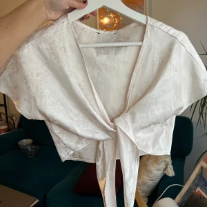 Vit blommig knytblus - Säljer en elegant vit/rosa blus med blommigt mönster och knytning framtill. Blusen har korta ärmar och en vacker glansig finish som ger en lyxig känsla. Perfekt för att lyfta vilken outfit som helst!