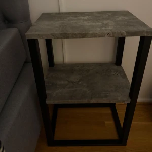 Grått sidobord i betongstil - Snyggt och modernt sidobord i grå betongstil med två hyllplan. Bordet har en robust metallram i svart som ger en industriell känsla. Perfekt som nattduksbord eller för att ställa dekorativa föremål på. Passar bra i både sovrum och vardagsrum. En är lite skadad men inte alls mycket 