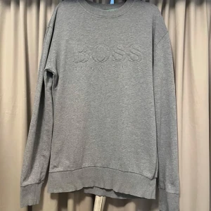 Grå sweatshirt från Hugo Boss - Säljer en stilren grå sweatshirt från Hugo Boss med broderad logga på bröstet. Tröjan har långa ärmar och ribbade muddar vid ärmslut och nederkant. Perfekt för en avslappnad look. Tröjan är insydd på sidorna ( hos skräddare ). Passar någon som är lång och smal. Axelbredd 51, Längd 68 