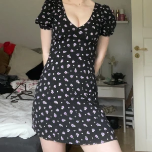 Blommig klännig - Säljer en söt svart klänning med lila blommor från H&M Divided. Klänningen har korta puffärmar och en v-ringad hals. Den har en knytning i ryggen och dragkedja för enkel på sättning. Använd få gånger❤️🩷Passar en Xs också eftersom det går att själv välja hur tajt den ska vara i midjan🩵💞