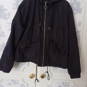 Svart vindjacka med dragkedja - Snygg svart vindjacka med dragkedja och justerbar huva. Jackan har praktiska fickor framtill och dragsko i nederkant för en anpassad passform. Perfekt för blåsiga dagar.