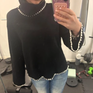 Svart stickad polotröja - Säljer en svart stickad polotröja med vita detaljer vid krage, ärmslut och nederkant. Tröjan har långa ärmar och en mysig passform. Perfekt för kyliga dagar!