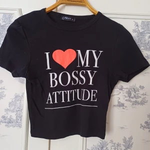 Svart croppad t-shirt från FB Sister - Säljer en svart croppad t-shirt från FB Sister med texten 'I ❤️ MY BOSSY ATTITUDE' i vitt och rött. Perfekt för att uttrycka din attityd med stil. Kortärmad och bekväm passform.