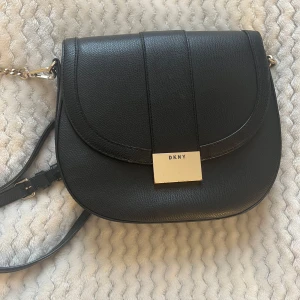 Svart axelväska från DKNY - Snygg svart axelväska från DKNY i skinn med en elegant design. Väskan har en justerbar axelrem med kedjedetaljer och en stilren metallplatta med logotyp på framsidan.