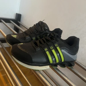 Svarta Adidas sneakers med gröna detaljer - Snygga svarta Adidas sneakers med gröna detaljer och snörning. Skorna har en unik sula med grön och vit färg. Perfekta för en sportig look.