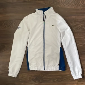 Vit windbreaker från Lacoste - Säljer en stilren vit vindjacka från Lacoste med dragkedja och det klassiska krokodilmärket på bröstet. Den är aldrig använd och i nyskick. 