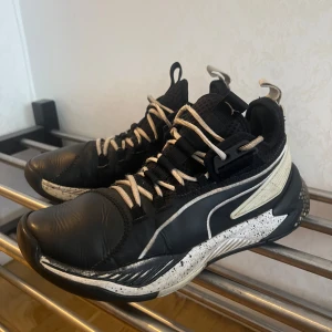 Svarta och vita sneakers från Puma - Snygga svarta och vita sneakers från Puma med en unik design. Skorna har snörning och en robust sula med prickigt mönster. Perfekt för basket.