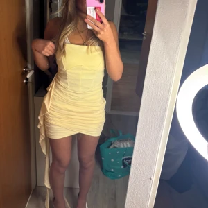 Ljusgul kortklänning - Snygg ljusgul kortklänning med en tight passform och volangdetaljer på sidan. Klänningen är ärmlös och har en stilren design som framhäver figuren. Perfekt för sommarens festligheter!