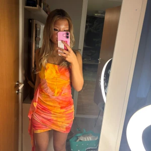 Färgglad klänning med omlott - Säljer en färgglad klänning i orange och rosa toner. Klänningen är axelbandslös och har en omlott-design med en snygg drapering på sidan. Perfekt för sommarens fester!