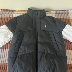 Svart och vit pufferjacka från Calvin Klein - Säljer en snygg svart och vit pufferjacka från Calvin Klein Jeans. Jackan har en dragkedja framtill och är långärmad med ett stilrent märke på bröstet. Väldigt bra för kyliga dagar när du vill hålla stilen.