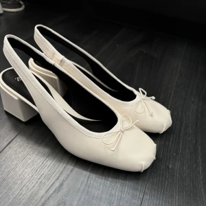 Vita pumps - Stilrena vita pumps från Zara med en liten rosett framtill. Skorna har en fyrkantig klack och öppen häl, vilket ger en elegant touch. Perfekta för en chic look. Aldrig använda 