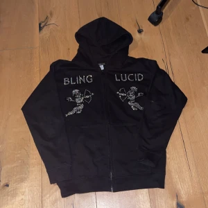 Svart hoodie med blingdetaljer - Svart hoodie med dragkedja och huva. Framsidan pryds av texten 'BLING LUCID' och två änglar i blingdetaljer. Perfekt för en cool och avslappnad stil. Storlek L. Använd några gånger men ser inget fel.