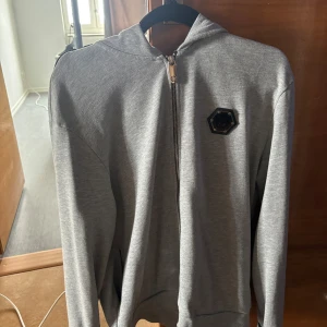 Grå hoodie från Philipp Plein - Grå hoodie från Philipp Plein med dragkedja och logga på bröstet och ryggen. Det är i storlek XL MEN passar M/L (1000kr)