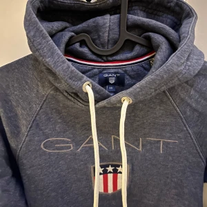 Mellanblå  hoodie från GANT - Säljer en snygg mellanblå  hoodie från GANT med logga och emblem på bröstet. Tröjan har en justerbar huva med vita snören. Inte mycket använd.
