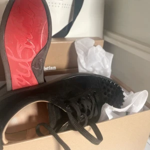 Skor  - Säljer ett par svarta sneakers  med klassisk röd sula och svarta nitar på tån. Skorna har snörning och är tillverkade i mocka. Perfekta för dig som vill sticka ut med en lyxig och edgy stil.