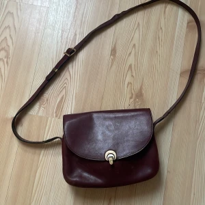 Vinröd axelväska från Etienne Aigner - Säljer en klassisk vinröd axelväska i skinn från Etienne Aigner med justerbar rem och snyggt metallspänne framtill. Väskan har ett stort huvudfack och innerficka med dragkedja. Perfekt för dig som gillar stilrena accessoarer. Remen hade behövts sys igen då tråden släppts, sen har den lite färg på insidan(syns bara om man öppnar)