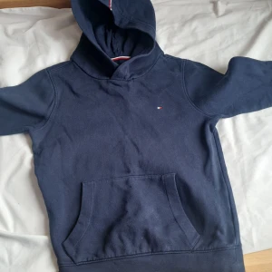 Marinblå hoodie från Tommy Hilfiger - Säljer en marinblå hoodie från Tommy Hilfiger med klassisk logga på bröstet och tryck på huvan. Tröjan har känguruficka och ribbade muddar. Perfekt för en avslappnad stil.