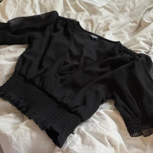 Svart blus med v-ringning från Bubbleroom - Säljer en svart blus från Bubbleroom med v-ringning och smockad midja samt ärmslut. Blusen har korta puffärmar och volangdetaljer vid midjan och ärmsluten. 