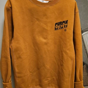 Mustardgul sweatshirt från Purple Velvet Eytys - Säljer en snygg mustardgul sweatshirt från Purple Velvet Eytys med svart tryck på bröstet. Tröjan har rund halsringning och långa ärmar. Perfekt för dig som gillar streetwear och vill ha en bekväm och stilren look. Passar till både man och kvinna.