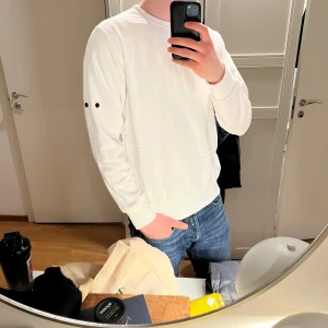 Vit sweatshirt från Stone Island - Säljer en stilren vit sweatshirt från Stone Island. Tröjan har rund halsringning, långa ärmar och en diskret svart knappdetalj på ena ärmen. Perfekt för en clean och enkel look. *BADGE SAKNAS*