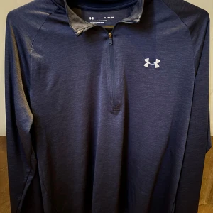 Mörkblå långärmad träningströja från Under Armour - Säljer en mörkblå långärmad träningströja från Under Armour med halv dragkedja och logga på bröstet. Tröjan har en loose passform och är perfekt för träning eller sportaktiviteter.