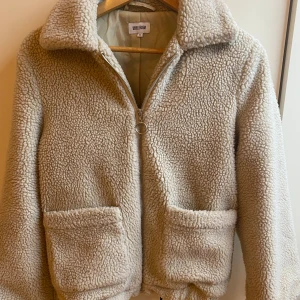 Beige teddyjacka från Bubbleroom - Mysig beige teddyjacka från Bubbleroom med dragkedja och stora fickor framtill. Perfekt för kyliga dagar med sin fluffiga och varma design. Jackan har en klassisk krage och är långärmad.
