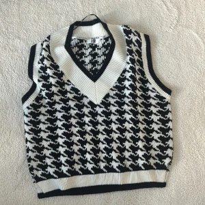 Svartvit stickad väst från Joy - Snygg svartvit stickad väst från Joy med ett klassiskt hundtandsmönster. Västen har en ribbad v-ringning och är perfekt för lager-på-lager-stil. Ett stilrent plagg som passar till många olika outfits.