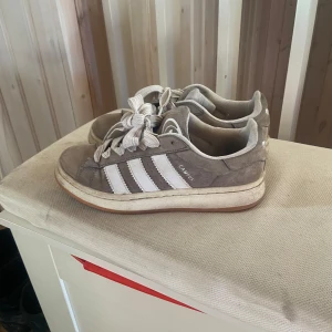 Grå Adidas Campus sneakers - Säljer ett par grå Adidas Campus sneakers med vita ränder och snörning. Skorna har en klassisk design med rund tå och är perfekta för vardagsbruk. Skoinlägget  kan behövas byta då den är lite sliten.