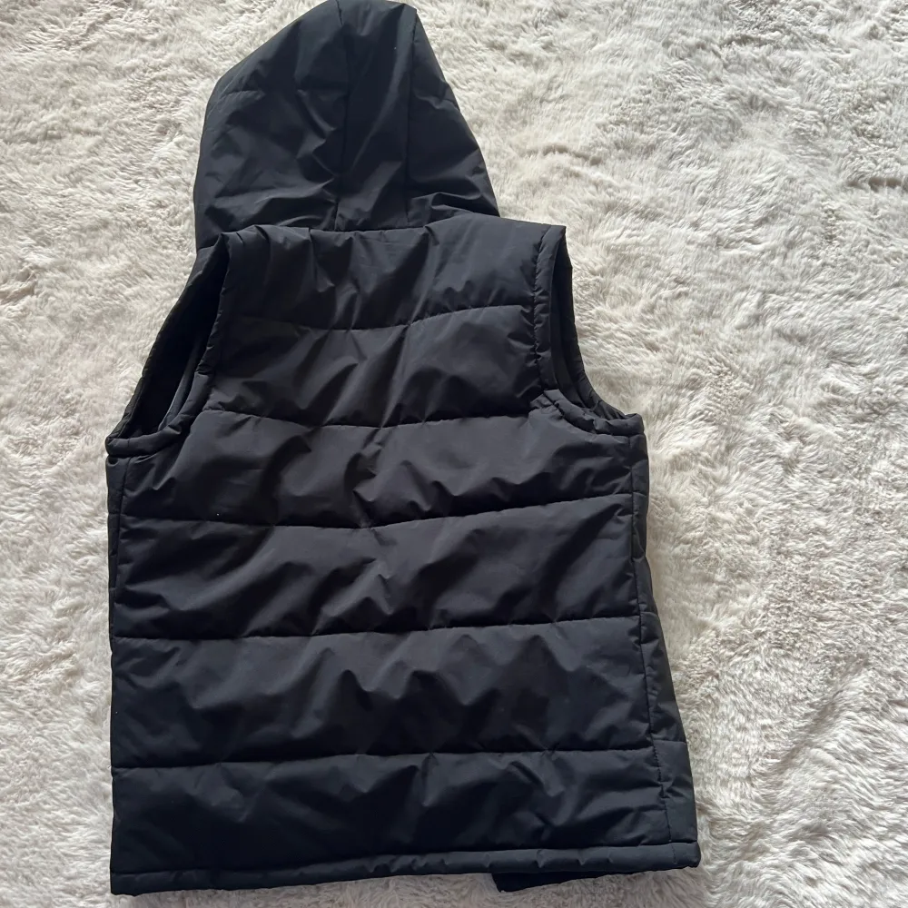 Snygg svart dunväst med dragkedja och praktiska fickor. Perfekt för kyliga dagar med sin quiltade design och huva som håller dig varm. En stilren och funktionell väst för alla tillfällen.. Takit.