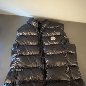 Svart dunväst från Moncler - Snygg svart dunväst från Moncler med glansig yta och dragkedja framtill. Västen har en broderad logotyp på bröstet och är perfekt för kyligare dagar. Den är både stilren och funktionell. Pris kan absolut diskuteras!