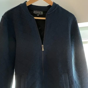 Mörkblå quiltad jacka från Sir Raymond Tailor - Snygg mörkblå jacka från Sir Raymond Tailor med quiltat mönster. Jackan har en dragkedja framtill och är långärmad. Perfekt för en stilren look. Aldrig använd 10/10 skick! 