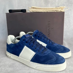 Valentino flycrews - Säljer ett par snygga Valentino Garavani sneakers i blå mocka med vita detaljer och vit sula. Skorna har rund tå, snörning och ikoniska nitar på hälen. Perfekta för dig som vill ha något unikt och stilrent, storlek 43