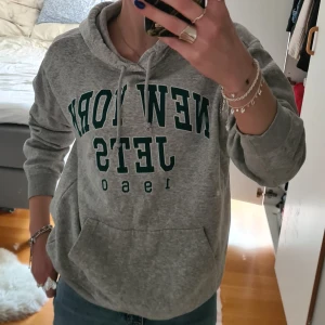 Grå hoodie med New York Jets-tryck - Säljer en grå hoodie med grön text 'New York Jets 1960' på bröstet. Tröjan har huva med snören och en stor magficka framtill. Perfekt för en sportig och avslappnad stil. Sälj för använder it längre💕