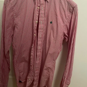 Röd/rosa- och vitrandig skjorta från Morris - Säljer en klassisk röd- och vitrandig skjorta från Morris med lång ärm och knappar framtill. Skjortan har en liten grön broderad detalj på bröstet och är perfekt för dig som gillar stilrena plagg med lite färg.