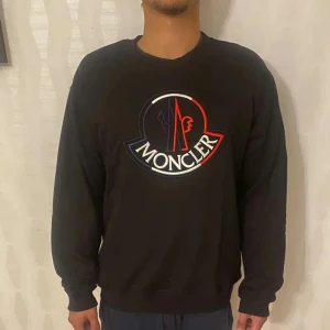 Svart långärmad tröja från Moncler - Säljer en stilren svart långärmad tröja från Moncler med ett stort logotyptryck på bröstet i rött, vitt och blått. Tröjan har en rund halsringning och Moncler-märke på ärmen. Perfekt för en casual look.