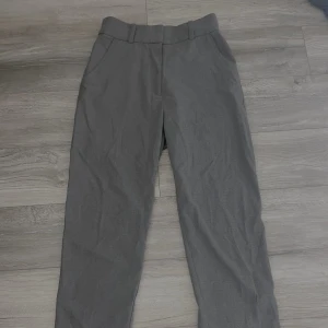 Grå kostymbyxor - Snygga grå kostymbyxor med hög midja och rak passform. Perfekta för en stilren look. Byxorna har en klassisk design med hällor för bälte och diskreta fickor baktill.