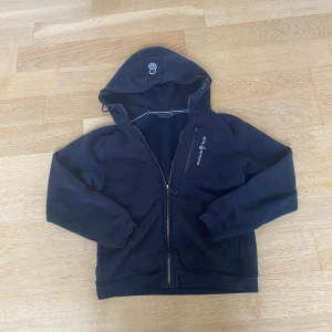 Svart hoodie från Sail Racing - Säljer en svart hoodie från Sail Racing med dragkedja och broderad logga på bröstet. Den har en praktisk huva och långa ärmar. Perfekt för en casual look eller för att hålla sig varm under kyliga dagar.