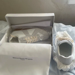 Maison Margiela Skor - Snygga vita sneakers från Maison Martin Margiela med gråa mockadetaljer och snörning. Skorna har en klassisk design med rund tå och en brun sula som ger en stilren look. Perfekta för en trendig vardagsstil. Dom är helt nya och äkta. 