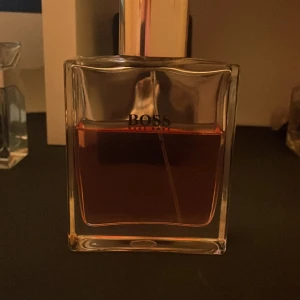 Parfym från Boss - Elegant parfym från Boss i en stilren, fyrkantig flaska med en gyllene vätska. Flaskan har en silverfärgad kork och en minimalistisk design som utstrålar lyx och klass. Perfekt för den som vill ha en sofistikerad doft.