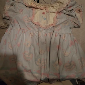 Pastellfärgad lolita klänning med volangdetaljer - Charmig klänning i pastellfärger med ett lekfullt mönster av ansikten och text. Klänningen har volangdetaljer vid kragen och korta ärmar. Perfekt för den som gillar en söt och unik stil.