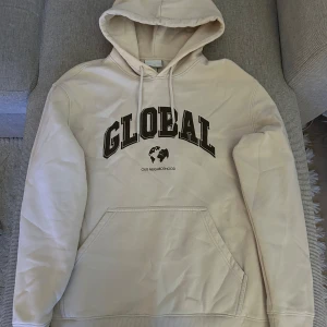 Beige hoodie med tryck från H&M - Säljer en beige hoodie från H&M med svart text 'GLOBAL' och jordglobstryck på bröstet. Tröjan har huva med dragsko och en stor magficka. Perfekt för en avslappnad stil.