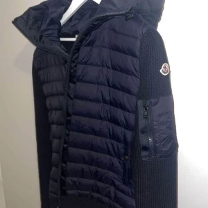 Moncler cardigan  - Snygg mörkblå pufferjacka från Moncler med stickade ärmar och huva. Jackan har dragkedja framtill, en ficka med dragkedja på ärmen och quiltad framsida. Perfekt för kyliga dagar och ger en sportig look.storlek S